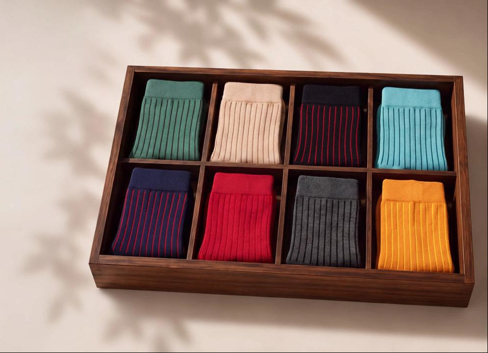 A wooden box with eight compartments holding folded ribbed socks in multiple colors, arranged on a light beige surface with soft leaf shadows.